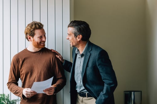 Deux hommes souriants en pleine discussion concernant un document écrit, probablement un contrat de location, dans un intérieur lumineux avec des fenêtres à lamelles verticales : le plus jeune, vêtu d'un pull-over marron, tient une feuille de papier tandis que l'autre, habillé d'un costume gris avec chemise, pose une main rassurante sur son épaule, illustrant un échange bienveillant entre un propriétaire et un futur locataire évoquant les garanties nécessaires pour une location.