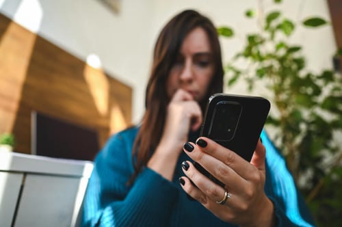 Jeune femme propriétaire ou locataire en intérieur moderne vérifiant attentivement des informations sur son smartphone, expression réfléchie et main posée sur le menton, illustrant la vigilance nécessaire pour détecter les faux garants et faux dossiers dans le cadre d'une location immobilière.