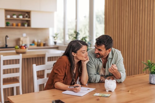 Couple calculant leur taux d’endettement maximum autour d’une table en bois dans une cuisine moderne et lumineuse. La femme, concentrée, prend des notes sur un carnet avec un stylo tandis que l’homme, souriant, explique des détails financiers en tenant des documents et une calculatrice. Sur la table, un petit cochon tirelire blanc et des pièces de monnaie symbolisent l’épargne et la gestion budgétaire. En arrière-plan, une cuisine bien équipée avec des étagères, des plantes et une fenêtre laissant entrer la lumière naturelle. Illustration parfaite pour comprendre comment évaluer son taux d’endettement avant un emprunt immobilier ou un crédit.