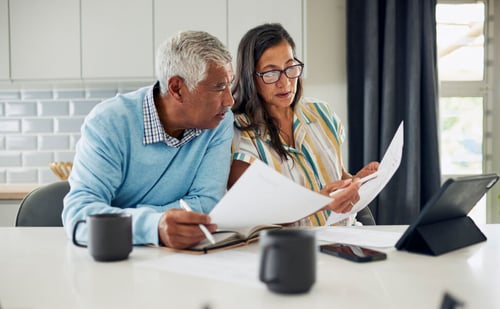 Un couple senior étudiant attentivement des documents fiscaux assis à une table avec des mugs et un tableau numérique, illustrant la préparation du calcul du taux d'imposition pour une location meublée, dans un cadre domestique éclairé et organisé. L'homme, vêtu d'un pull bleu clair, prend des notes dans un carnet tandis que la femme, portant des lunettes et une chemise rayée, consulte des papiers officiels, reflétant une démarche sérieuse de gestion fiscale et de compréhension des obligations liées à la fiscalité locative.