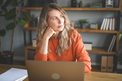 Une propriétaire ou gestionnaire immobilière réfléchit attentivement devant son ordinateur portable dans un bureau aménagé, avec des étagères remplies de dossiers et de livres en arrière-plan. Habillée d’un pull orange et les cheveux longs et blonds, elle semble analyser une situation complexe, illustrant les questionnements juridiques liés à la sélection des dossiers de location et aux risques de poursuites.