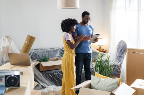 Deux individus analysant des documents ensemble dans un appartement en cours de déménagement, avec un homme et une en tenant un appareil numérique, entourés d’objets de déménagement et de cartons, avec un fond de pièces partiellement emballées et des meubles protégés par des housses, évoquant la gestion des responsabilités liées à un logement, dans un contexte de transition résidentielle, rappelant l’importance de bien comprendre les étapes de gestion d’un habitat, incluant la résiliation d’assurance habitation.