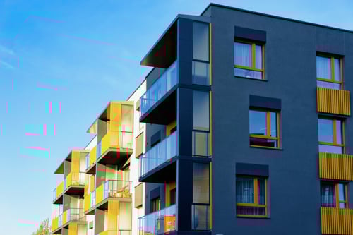 Immeuble moderne de plusieurs étages avec balcon vitré et garde-corps, illustrant un logement locatif récent : façades aux tons gris et jaunes, fenêtres à encadrements orange et grands espaces extérieurs privatifs, idéal pour aborder les travaux et aménagements possibles par un propriétaire pendant une période de location.