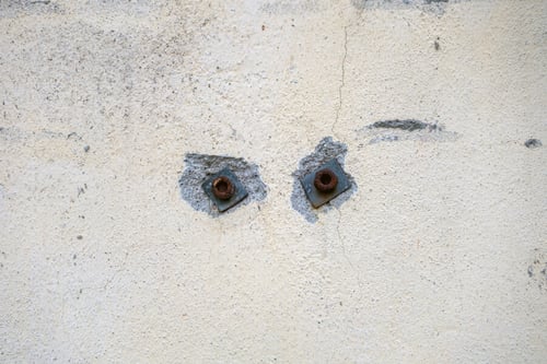 Deux fixations murales rouillées avec écrous encore en place dans un mur enduite de plâtre fissuré et dégradé, illustrant les traces de trous laissés après le retrait d'étagères ou d'éléments fixés au mur lors d'un état des lieux de sortie.