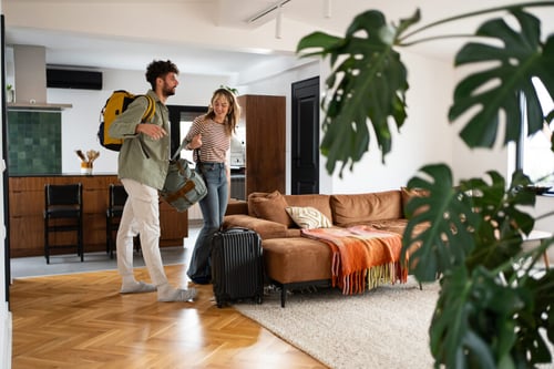 Deux personnes souriantes préparant un voyage dans un appartement meublé lumineux, illustrant l'impact des modifications de la TVA sur les locations touristiques en 2026. L’homme porte un sac à dos jaune et tient une valise, tandis que la femme, avec une valise noire à roulettes, semble discuter joyeusement. Le salon moderne comprend un canapé en velours marron, des coussins décoratifs, une couverture tricotée, une table basse et des plantes d’intérieur. En arrière-plan, une cuisine équipée et des meubles en bois ajoutent au charme de ce logement de vacances, sujet aux nouvelles réglementations fiscales concernant la TVA pour les locations saisonnières.