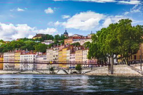 Vue panoramique des berges de la Saône à Lyon par une journée ensoleillée, mettant en avant les façades colorées et typiques des immeubles du Vieux Lyon, avec leurs tons pastel allant du jaune au rose en passant par l'orange. Au premier plan, les eaux calmes de la rivière reflètent partiellement les bâtiments historiques, tandis qu'un pont piéton suspendu relie les deux rives. En arrière-plan, la colline verdoyante de Fourvière domine le paysage avec ses arbres et ses habitations, soulignant le charme architectural et géographique de Lyon, ville au cœur des débats sur l'encadrement des loyers et son impact sur le marché immobilier local.