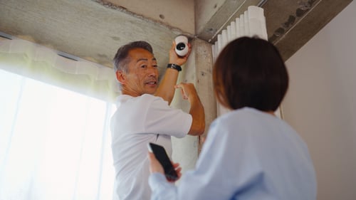 Homme installant une caméra de vidéosurveillance dans le coin supérieur d une pièce en intérieur, aidé par une femme tenant un smartphone, idéal pour illustrer les étapes d installation d un système de sécurité en location. Scène montrant les détails pratiques de la pose d une caméra au plafond près d un mur, avec des câbles apparents et des accessoires de fixation visibles.