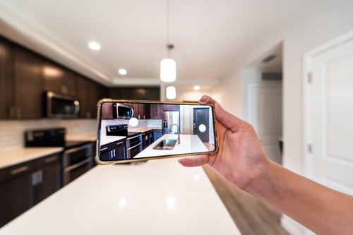 Main tenant un smartphone affichant une visite virtuelle immersive d'une cuisine moderne avec superposition de l'image réelle en arrière-plan, montrant des meubles en bois foncé, un plan de travail blanc et des appareils électroménagers encastrés, idéale pour illustrer les avantages des visites virtuelles pour les propriétaires souhaitant mettre en valeur leur bien immobilier.