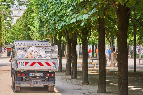 Véhicule utilitaire de collecte des déchets en action dans un parc urbain arboré, chargé de sacs et cartons, avec une signalétique indiquant une limitation de vitesse à 40 km/h, licence française visible GH-237-VD. Scène illustrant la gestion des ordures ménagères en milieu public, avec des passants en arrière-plan, idéale pour évoquer les enjeux de la taxe des ordures ménagères et leur collecte en ville.