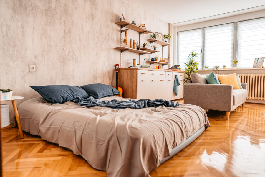 Découvrez un intérieur moderne et cosy idéal pour une location courte durée conforme aux nouvelles réglementations de l'UE sur les locations Airbnb. Cet espace lumineux et bien agencé comprend un grand lit confortable avec une literie neutre et des coussins bleus, parfait pour attirer les voyageurs en quête de confort. À proximité, un salon accueillant avec canapé et fauteuils assortis de coussins colorés offre un espace détente. La cuisine ouverte, équipée d'étagères en bois et d'accessoires pratiques, répond aux attentes des locataires recherchant des commodités fonctionnelles. Les grandes fenêtres laissent entrer une abondante lumière naturelle, tandis que le parquet en bois ajoute une touche chaleureuse. Parfait pour optimiser vos annonces de location courte durée en respectant les nouvelles règles européennes pour les plateformes comme Airbnb.
