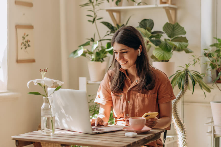Femme souriante travaillant sur un ordinateur portable dans un intérieur lumineux et cosy, illustrant le télétravail depuis un logement en location. Elle porte un chemisier en jean tout en tenant un carnet de notes et une tasse de café posée sur une table en bois. Autour d’elle, des plantes vertes en pots et une décoration soignée créent un cadre de travail agréable et productif. Cette image représente la possibilité d’exercer une activité professionnelle à domicile, même en location, en optimisant l’espace et le confort pour le travail à distance. Idéale pour évoquer les solutions pratiques et légales du télétravail en location.