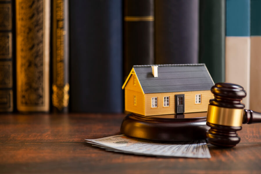 Petit modèle de maison jaune posé sur un bureau en bois devant un marteau de juge et des billets de banque symbolisant un litige immobilier. Cette image illustre les risques de surestimation immobilière pouvant mener à des poursuites judiciaires pour un agent immobilier. Contexte juridique et financier lié aux conflits entre vendeurs, acheteurs et professionnels de l'immobilier lors d'une évaluation erronée de la valeur d'un bien. Idéale pour un article sur les conséquences légales et les recours en cas de surévaluation immobilière.