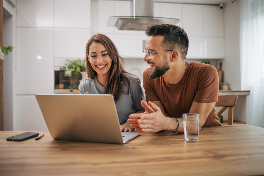 Jeune couple souriant consultant des informations sur un ordinateur portable dans une cuisine moderne et lumineuse. Les deux personnes semblent discuter avec enthousiasme des démarches administratives liées aux aides au logement. L’homme et la femme, assis à une table en bois, examinent ensemble des données en ligne, probablement pour vérifier leur éligibilité aux aides financières comme les APL ou autres dispositifs d’aide au logement en 2026. Ambiance conviviale et studieuse reflétant la recherche d’informations pratiques pour les locataires. Contexte adapté à un article sur les critères d’éligibilité aux aides au logement.