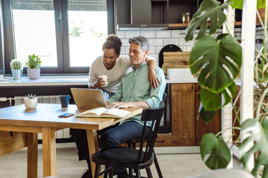 Deux personnes discutent des options d’assurance habitation pour locataires dans une cuisine moderne et lumineuse. L’homme, assis à une table en bois avec un ordinateur portable ouvert devant lui, consulte des documents tout en souriant. La femme, debout à ses côtés, pointe un détail sur l’écran tout en tenant une tasse de café. La pièce est décorée avec des plantes vertes, des pots sur le rebord de la fenêtre et une étagère en bois, créant une ambiance chaleureuse et organisée. Cette scène illustre la recherche d’une assurance locataire adaptée, combinant sécurité et tranquillité d’esprit dans un cadre de vie agréable. Idéal pour comprendre les critères de choix d’une assurance habitation pour locataires.