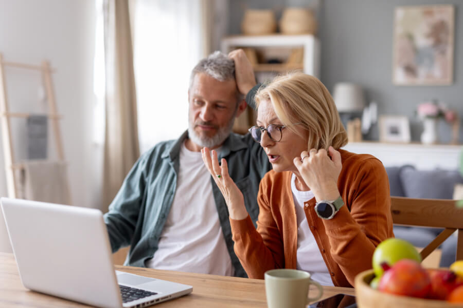 Deux seniors perplexes et inquiets consultent un site internet sur un ordinateur portable dans une cuisine moderne et lumineuse. L’homme, les mains sur la tête, semble stressé tandis que la femme, portant des lunettes, se tient la tempe en regardant l’écran avec une expression préoccupée. Cette scène illustre l’inquiétude liée à la découverte d’une arnaque en ligne, notamment l’arnaque aux loyers impayés ciblant les locataires. Les détails de leur environnement, comme les fruits sur la table et la décoration soignée, renforcent le réalisme de la situation et l’impact émotionnel de cette fraude financière sur des personnes vulnérables.