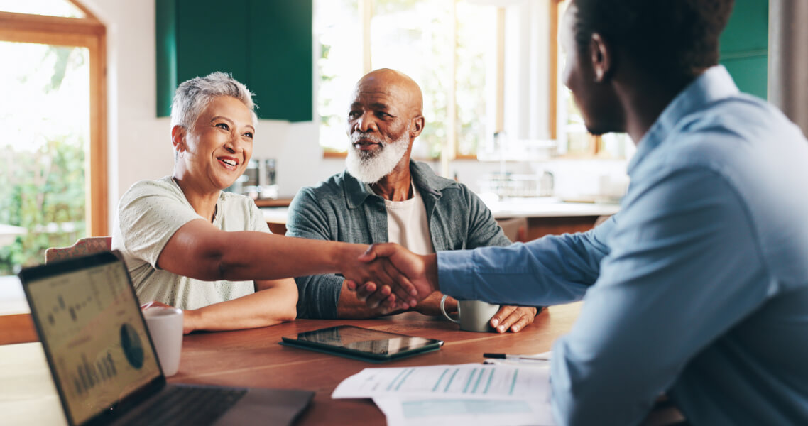Scène chaleureuse montrant un agent immobilier accompagnant un couple de seniors dans le financement de leur projet immobilier. Autour d'une table en bois dans un cadre lumineux et accueillant, l'agent, un homme souriant en chemise bleue, serre la main du couple âgé pour sceller un accord. Le senior, barbu et vêtu d'une veste grise, et sa compagne aux cheveux gris clair, habillée d'un haut clair, affichent des expressions de satisfaction et de confiance. Sur la table, des documents financiers, un contrat immobilier et une tablette affichant des graphiques illustrent les différentes étapes de l'accompagnement personnalisé. Cette image reflète l'importance d'un soutien expert pour les seniors souhaitant concrétiser leur projet immobilier en toute sérénité.