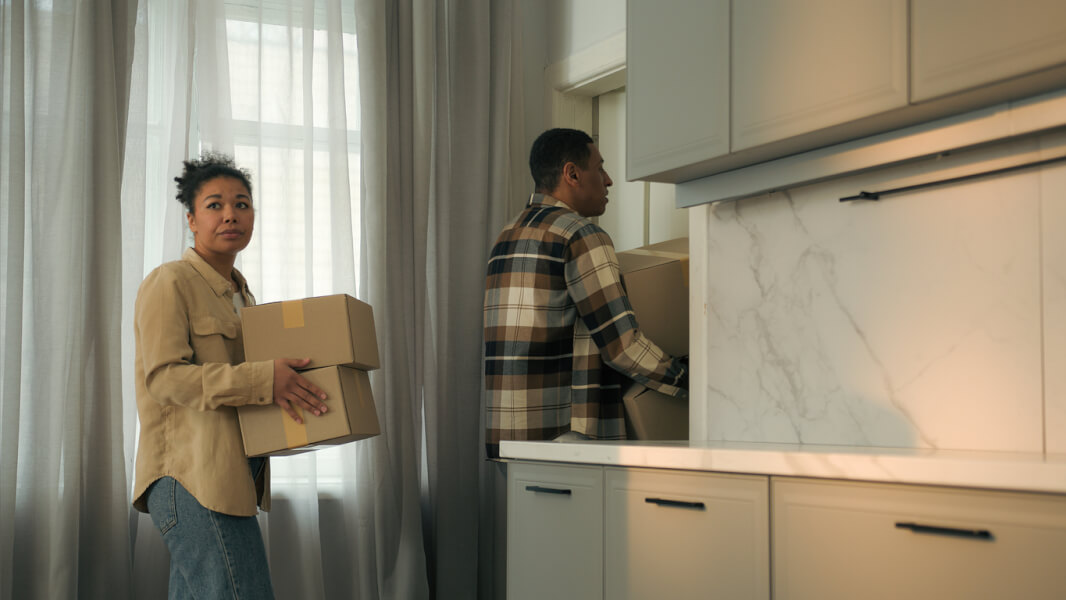 Deux personnes en train de déménager discutent dans une cuisine moderne après avoir déposé un préavis de départ. La femme, à gauche, tient un carton de déménagement marqué d’une étiquette tandis qu’elle semble poser une question ou échanger des informations. L’homme, à droite, place un autre carton sur un plan de travail de cuisine équipée de meubles clairs et d’un plan en marbre. Cette scène illustre une situation courante liée à la gestion d’un préavis de location et aux préparatifs de déménagement, notamment la possibilité de décaler la date de départ après le dépôt du préavis. Contexte idéal pour aborder les droits et démarches concernant la modification de la date de départ en fin de bail.