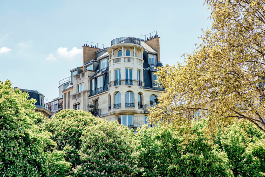 Découvrez un exemple typique de l'immobilier résidentiel en France illustrant la hausse des loyers analysée par l'Observatoire 2026. Cette photographie montre un immeuble haussmannien élégant à plusieurs étages, situé dans un quartier urbain probablement parisien. Les façades en pierre de taille, les balcons en fer forgé et les fenêtres hautes témoignent du charme architectural recherché, souvent associé à des loyers élevés. En premier plan, une végétation dense avec des arbres en fleurs souligne l’attrait des espaces verts dans les zones résidentielles prisées. Ce type de bien, emblématique des grandes villes françaises, est directement concerné par l’évolution des prix du marché locatif, un sujet central abordé dans notre analyse sur la hausse des loyers en France selon les dernières données de l'Observatoire 2026.