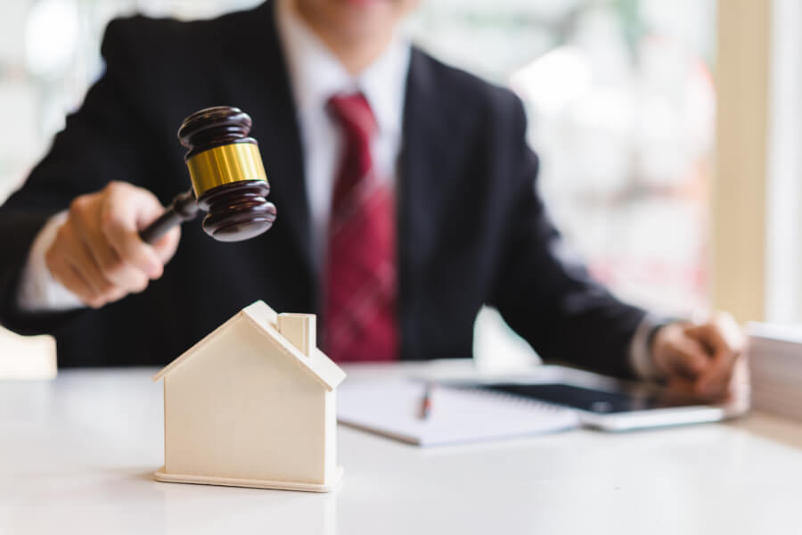 Juge en robe noire avec cravate rouge tenant un marteau de justice posé sur une maquette de maison blanche symbolisant une vente immobilière aux enchères judiciaires. Contexte de saisie immobilière et d'achat de bien saisi par décision de justice, illustrant les opportunités et procédures liées à l'acquisition d'un logement saisi à prix potentiellement avantageux. Idéal pour un article sur les avantages et précautions à prendre lors de l'achat d'un bien immobilier saisi.