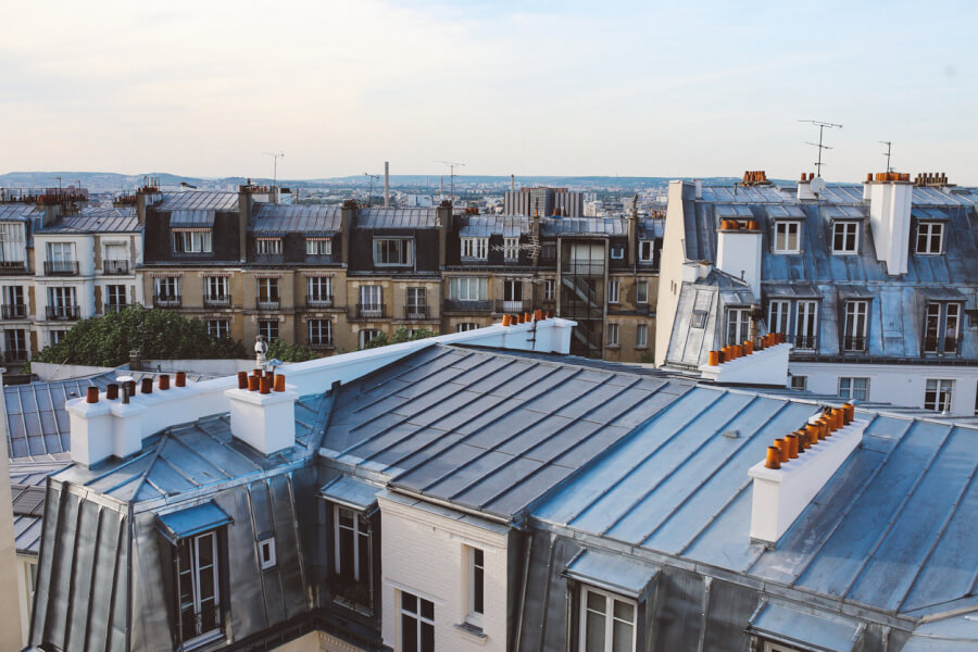 Vue panoramique des toits parisiens avec leurs caractéristiques architecturales typiques des immeubles haussmanniens en 2026. Cette image montre une diversité de toitures en zinc, certaines équipées de fenêtres de toit et de cheminées, offrant un aperçu des combles souvent aménageables en logements à Paris. Les bâtiments aux façades en pierre, avec leurs mansardes et leurs balcons en fer forgé, illustrent les opportunités de locations moins chères dans des quartiers moins centraux mais tout aussi charmants de la capitale. Louer un appartement sous les toits ou dans des espaces réaménagés peut représenter une solution économique pour réduire son budget logement à Paris en 2026, tout en profitant d’un cadre de vie authentique et pittoresque. Découvrez dans notre article des astuces pour dénicher ces pépites immobilières et optimiser votre recherche de location à moindre coût.