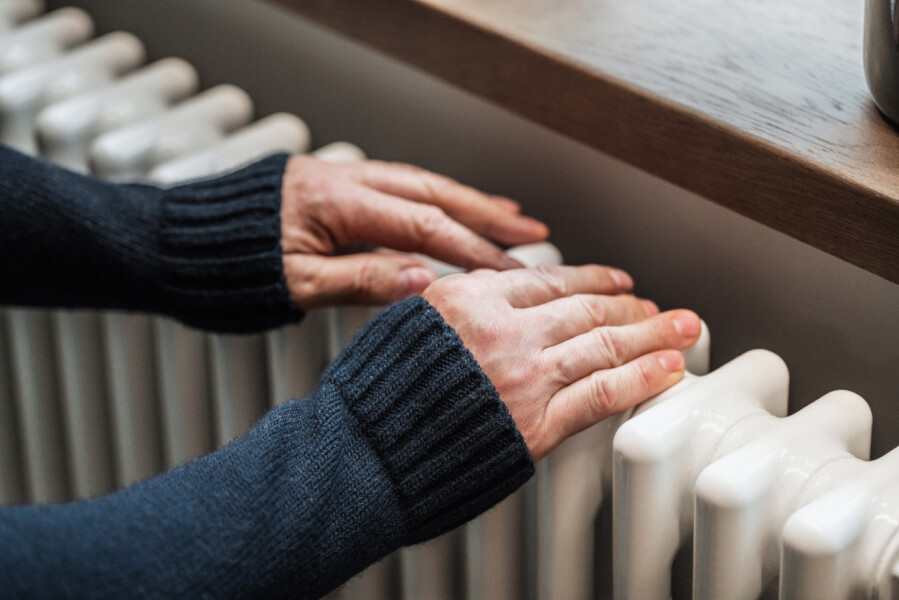 Une personne en pull chaud vérifie la température d'un radiateur ancien et peu efficace, illustrant le problème des passoires thermiques dans les logements mal isolés. Les mains posées sur un radiateur en fonte froid mettent en évidence l'inconfort thermique subi par les locataires en hiver. Cette image renvoie aux obligations légales des propriétaires concernant la rénovation énergétique et aux sanctions encourues en cas de non-respect des normes de performance thermique, notamment avec les nouvelles réglementations sur les logements énergivores. Contexte lié aux risques de gel des loyers, d'amendes ou d'interdictions de location pour les propriétaires négligeant la rénovation de leur passoire thermique.
