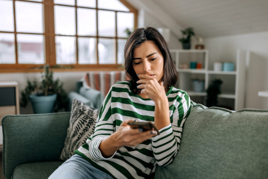 Femme inquiète assise sur un canapé vert dans un salon lumineux avec grandes fenêtres, consultant son téléphone portable avec un air préoccupé. Elle semble chercher des solutions en ligne concernant un propriétaire non réactif ou absent. Contexte illustrant les démarches à suivre lorsque un propriétaire ne répond plus aux locataires, incluant recherche d’informations juridiques ou conseils pratiques sur internet. Intérieur moderne et chaleureux avec étagères et plantes en arrière-plan. Idéal pour un article sur les recours en cas de silence du propriétaire.