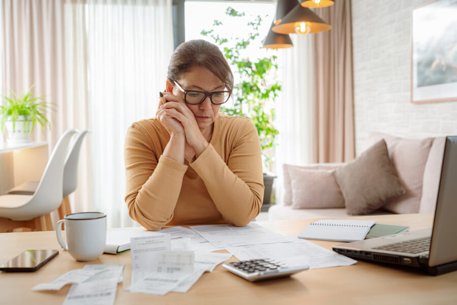 Femme stressée en train de gérer une dette locative et des problèmes de surendettement à domicile. Elle consulte des documents financiers, factures et relevés bancaires étalés sur une table en bois avec un ordinateur portable, une calculatrice et une tasse de café. La scène se déroule dans un salon lumineux et cosy, illustrant les difficultés financières et l'impact du surendettement sur les propriétaires. Cette image reflète les défis liés à la gestion des dettes locatives et les conséquences sur la situation personnelle et financière des propriétaires bailleurs. Idéale pour accompagner un article sur les solutions et recours en cas de surendettement immobilier.