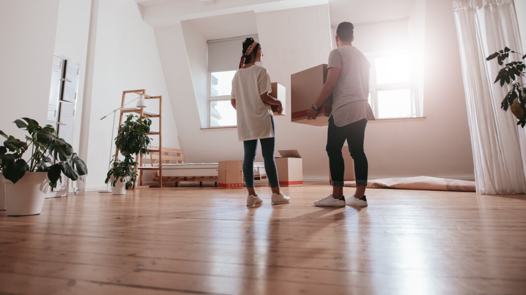Deux personnes en train de déménager dans un appartement lumineux et spacieux avec parquet, illustrant les implications pratiques du statut LMNP en 2026. La femme, debout près d’une fenêtre, discute avec l’homme qui porte un grand carton de déménagement. L’espace est partiellement meublé avec des étagères en bois, des plantes vertes en pots et quelques cartons empilés au sol. Cette scène reflète les préparatifs pour la location meublée non professionnelle, un sujet clé lié aux évolutions fiscales et réglementaires du statut LMNP dans le budget 2026. Idéal pour comprendre les avantages et les démarches de mise en location sous ce régime.