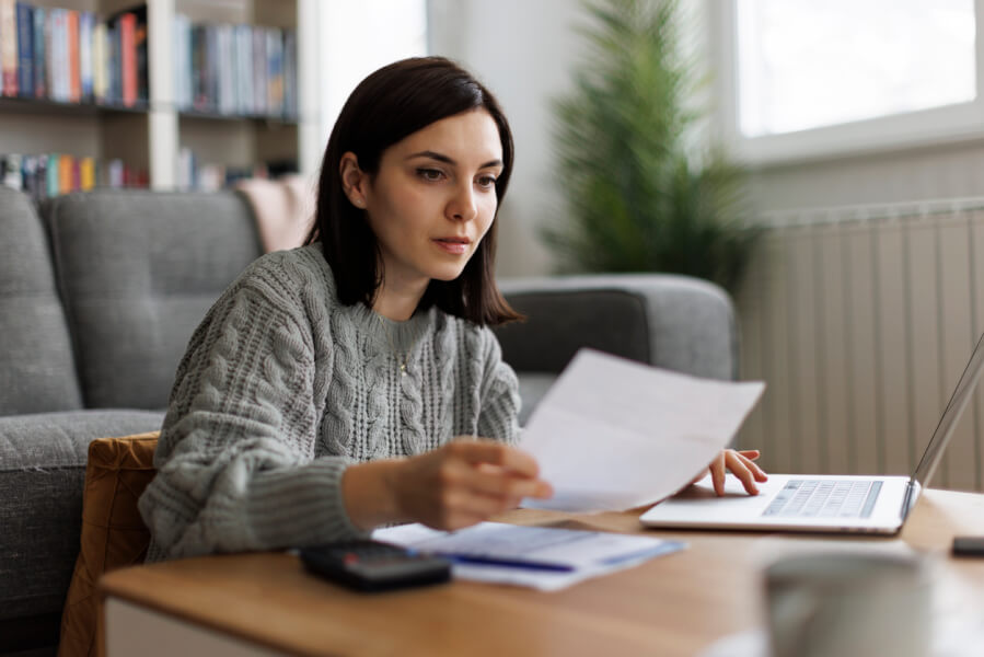 Jeune femme concentrée analysant les nouvelles dispositions du statut du bailleur privé pour le budget 2026 sur son ordinateur portable dans un salon moderne. Elle tient des documents imprimés détaillant les modifications légales et fiscales, avec un stylo et des notes à portée de main. À l’arrière-plan, une bibliothèque remplie de livres juridiques et financiers, illustrant la recherche et la préparation en matière de gestion locative et d’investissement immobilier. Contexte visuel adapté aux propriétaires bailleurs souhaitant comprendre les impacts des réformes fiscales à venir.