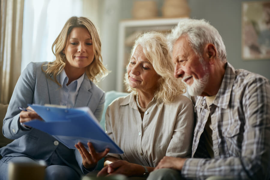 Une conseillère juridique ou notaire explique les détails d’une succession à un couple de personnes âgées, dans un cadre chaleureux et professionnel. Elle montre un document relatif à la gestion d’un bien immobilier loué, abordant spécifiquement la question de la récupération des loyers dans le cadre d’une succession. Cette scène illustre les démarches légales et les droits des héritiers concernant les revenus locatifs après le décès du propriétaire, en lien avec l’article Succession d’un bien loué : qui récupère les loyers.