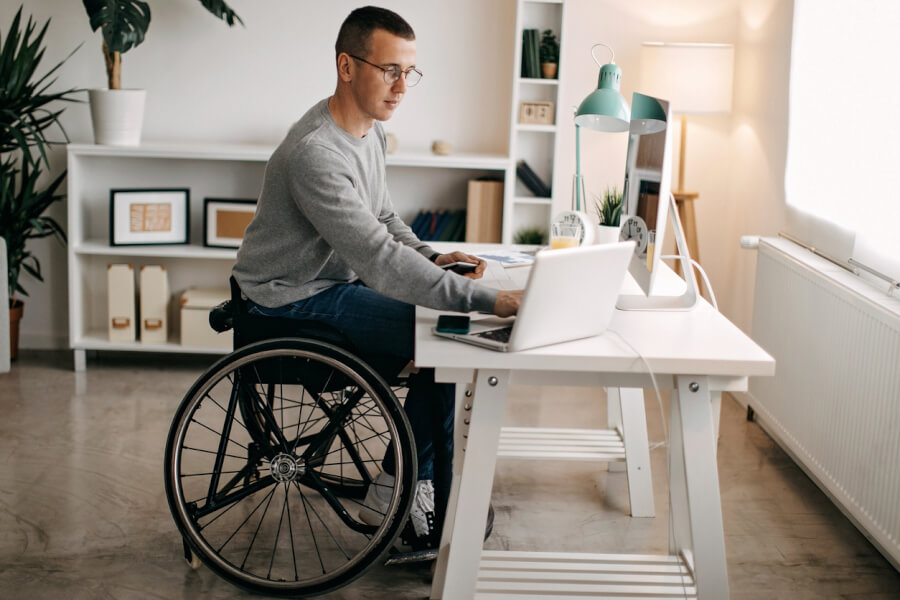 Homme en fauteuil roulant travaillant sur un ordinateur portable dans un bureau à domicile adapté. La pièce lumineuse et accessible est aménagée avec des étagères basses, un bureau ergonomique blanc et une lampe de travail verte. Ce visuel illustre l'importance d’un logement adapté pour les personnes en situation de handicap bénéficiant de l'Allocation aux Adultes Handicapés (AAH). L’image met en avant les solutions pratiques pour aménager un espace de télétravail ou de gestion administrative depuis chez soi, un critère essentiel pour les locataires handicapés recherchant un logement fonctionnel et accessible. Conseils pour trouver un logement avec l’AAH et optimiser son aménagement.