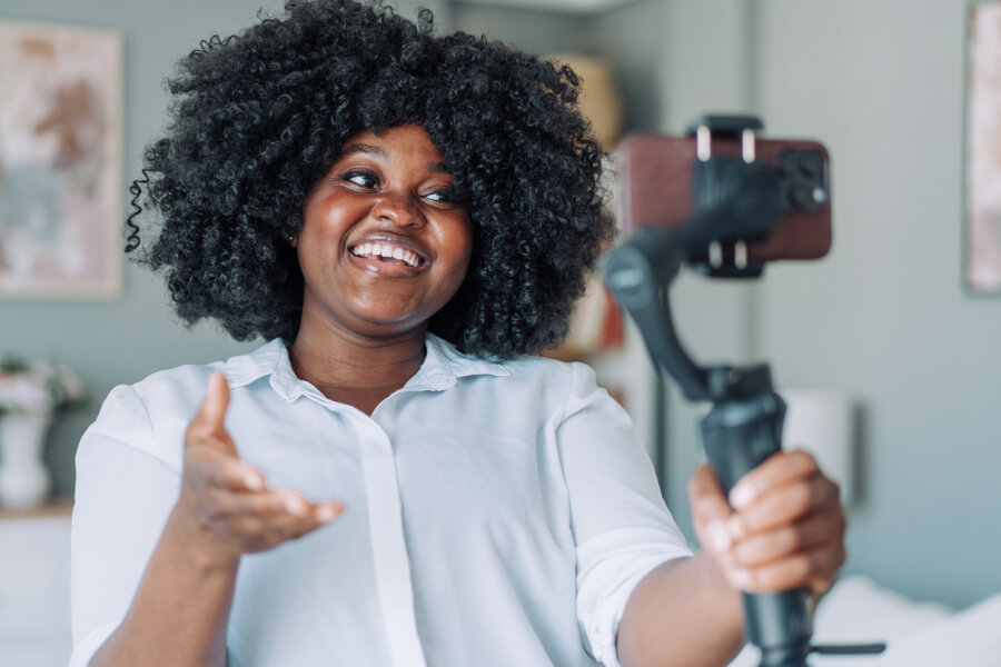 Jeune femme souriante et dynamique filmant une vidéo avec un smartphone monté sur stabilisateur gimbal dans un intérieur bien éclairé, idéale pour illustrer la création de contenus professionnels et attractifs afin de valoriser vos publications immobilières sur les réseaux sociaux.