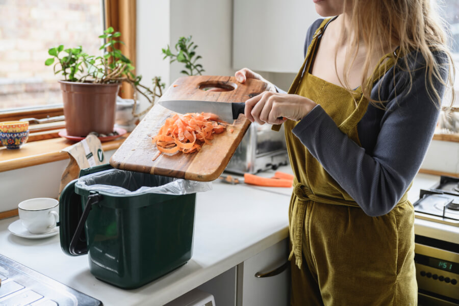 Une personne en tablier vert préparant des épluchures de carottes dans une cuisine bien éclairée et les déposant dans un seau de compost vert équipé d’un couvercle et d’un sac compostable, illustration concrète des gestes écologiques de compostage en intérieur adaptés aux logements en location.