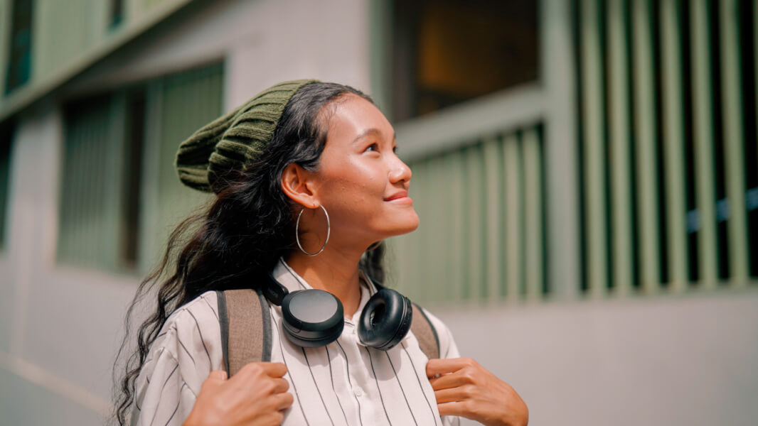 Jeune femme souriante et détendue en extérieur devant un bâtiment résidentiel avec des fenêtres vert pâle, portant des écouteurs autour du cou, un beanie vert et une veste striée, tenant les bretelles de son sac à dos, symbolisant la recherche de logement sereinement pendant une période d’essai professionnelle dans une nouvelle ville.