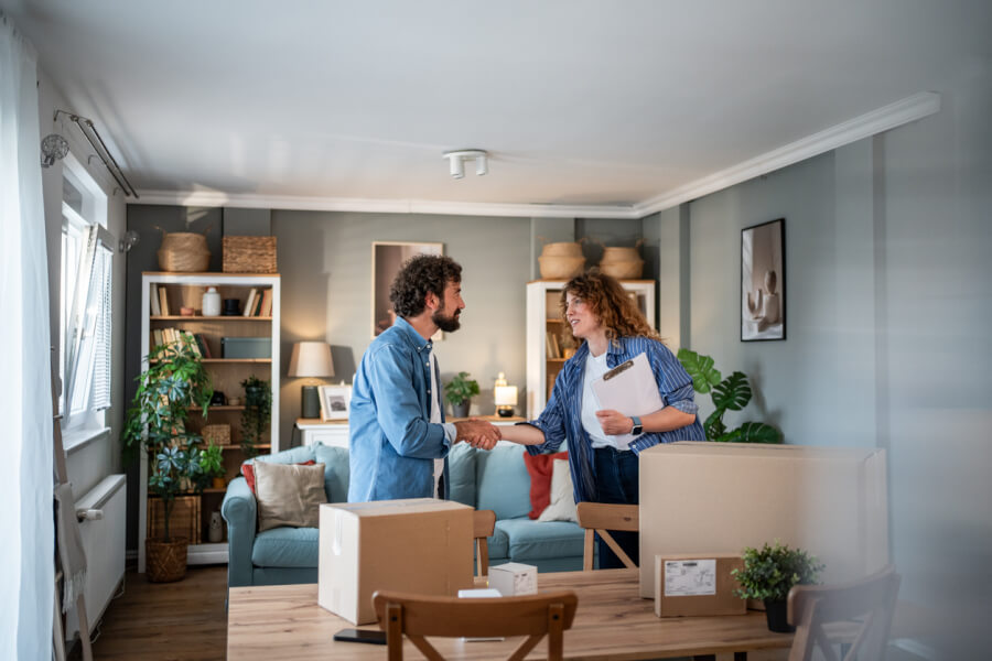 Un propriétaire et une locataire échangent une poignée de main amicale dans un salon lumineux lors de la remise des clés avant un départ de location anticipé. Autour d’eux, des cartons d’emballage et des plantes vertes décorent l’espace meublé avec un canapé bleu, des étagères remplies de livres et d’objets décoratifs, illustrant une transition organisée et sereine pour la fin de bail. La pièce, bien rangée et accueillante, reflète une gestion locative réussie et un état des lieux préparé avec soin.