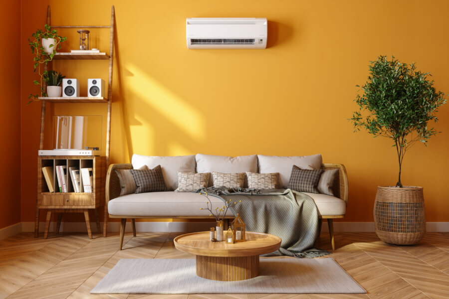 Découvrez un intérieur meublé moderne et chaleureux idéal pour comprendre l’encadrement des loyers des locations meublées. Ce salon lumineux aux murs orange clair est équipé d’un canapé confortable en tissu beige avec plusieurs coussins assortis, d’une couverture grise et d’une table basse en bois circulaire. À gauche, une étagère en bois élégamment organisée avec des plantes vertes, des livres, des enceintes et des éléments décoratifs. À droite, un grand pot en osier abrite une plante verte, ajoutant une touche naturelle à l’espace. Au-dessus du canapé, un climatiseur mural discret assure le confort thermique. Ce cadre illustre parfaitement les critères de décoration et d’équipement nécessaires pour une location meublée conforme aux réglementations sur l’encadrement des loyers. Parfait pour un logement étudiant ou une résidence temporaire, cet aménagement répond aux exigences légales tout en offrant un environnement accueillant.