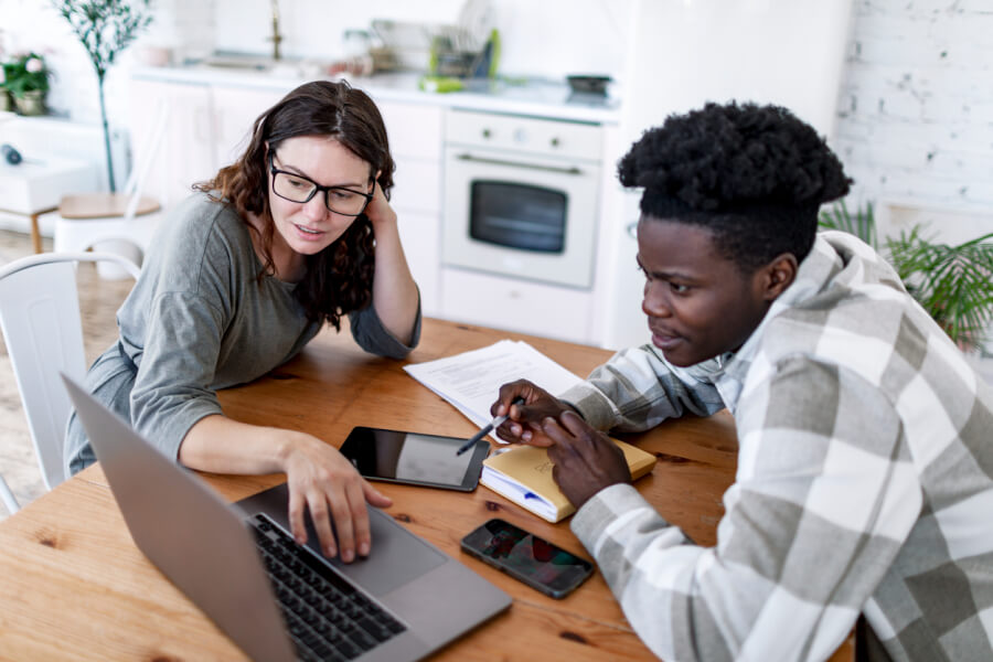 Deux personnes discutent des implications financières liées à la hausse de la taxe foncière prévue en 2026, assises autour d'une table en bois dans une cuisine moderne. La femme, portant des lunettes, consulte des informations sur un ordinateur portable tout en tenant un smartphone. À côté d'elle, un homme examine attentivement des documents papier et prend des notes avec un stylo. Sur la table sont également posés un deuxième smartphone et des papiers contenant des chiffres et des graphiques, illustrant une analyse détaillée des impacts économiques pour les propriétaires immobiliers. Cette scène reflète une préparation et une étude minutieuse des conséquences de la réforme fiscale.