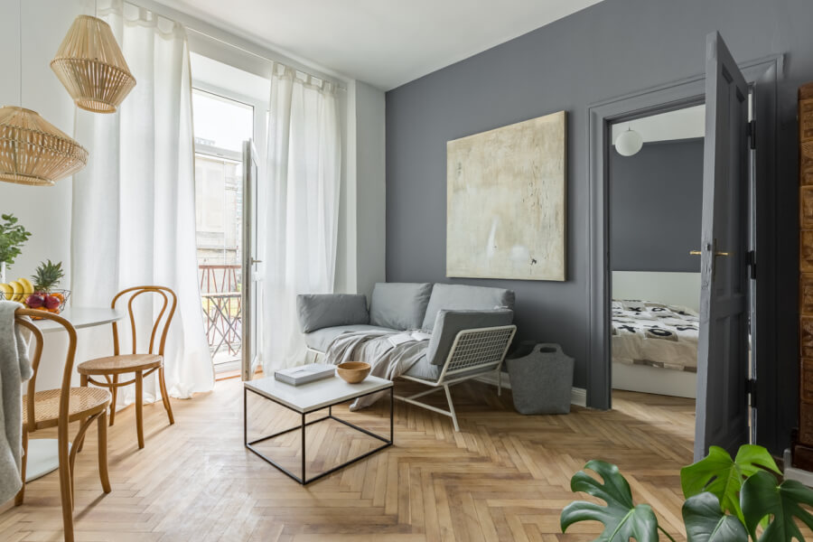 Description SEO optimisée pour texte alternatif : intérieur moderne d'un studio en location meublée éligible à la Cotisation Foncière des Entreprises (CFE) avec salon ouvert sur balcon. Pièce lumineuse avec sol en parquet clair en motif chevron, murs gris anthracite et grande baie vitrée donnant sur un petit balcon. Mobilier fonctionnel incluant un canapé-lit gris avec coussins, une table basse blanche à structure métallique, et deux chaises en bois style bistrot autour d'une table ronde. Éclairage design avec suspension en osier naturel et lampe murale. Espace optimisé avec porte ouverte sur une chambre équipée d'un lit et rangements. Décoration épurée avec une grande toile abstraite au mur et plantes vertes pour une ambiance chaleureuse. Idéal pour illustrer les spécificités des logements meublés soumis à la CFE.