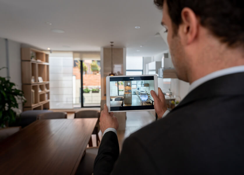 Homme en costume visualisant une visite virtuelle d’un appartement moderne sur une tablette tactile, illustrant la possibilité de découvrir un bien immobilier à distance sans se déplacer, avec en arrière-plan un intérieur contemporain comprenant un salon avec bibliothèque murale et table en bois.