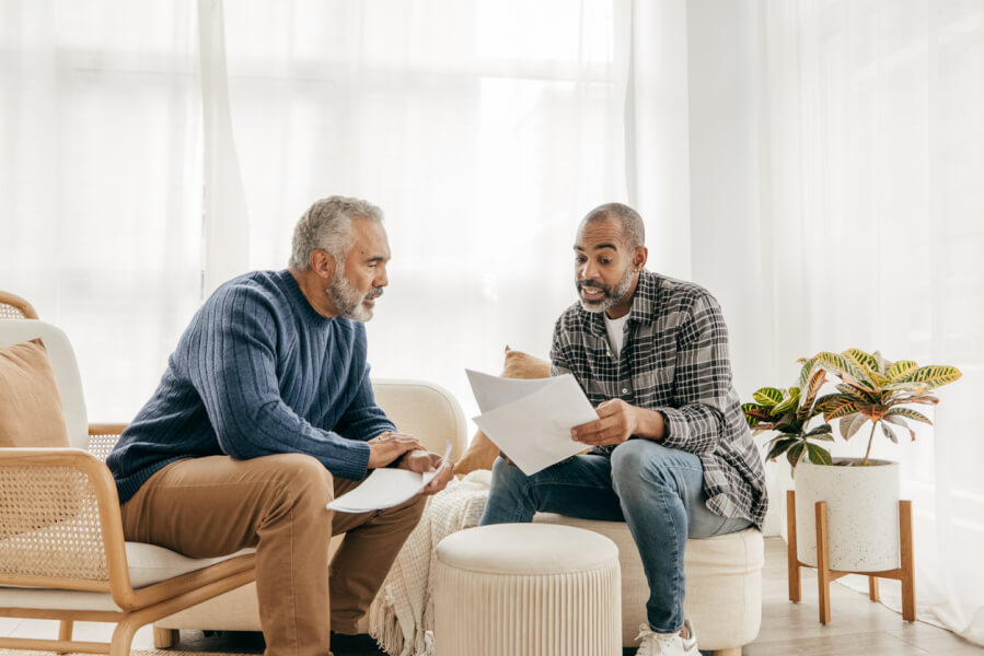 Deux hommes discutant de documents liés à la gestion locative dans un salon moderne et lumineux. L’un, assis sur une chaise en rotin avec un dossier à la main, semble examiner des papiers concernant un loyer. L’autre, installé sur un pouf, tient une feuille qui pourrait être un contrat de location ou une notification de révision de loyer. La scène illustre une situation où un propriétaire bailleur vérifie ou ajuste un loyer non révisé, entouré de mobilier contemporain et d’une plante verte, évoquant un cadre professionnel et serein pour aborder les questions de gestion immobilière et de régularisation des loyers.