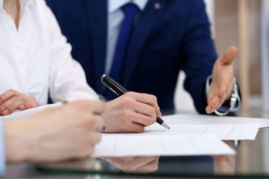Deux personnes en train de signer un contrat immobilier ou professionnel avec un agent en costume : une personne remplit et paraphe le document avec un stylo tandis qu'un professionnel en costume bleu, probablement un agent mandaté, tend la main pour finaliser l'accord, illustrant ainsi les interactions possibles dans le cadre d'un mandat d’entremise ou de représentation.
