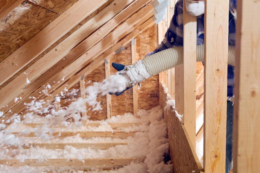 Technicien en train d'installer de la laine minérale en ouate dans les combles d'une maison pour améliorer l'isolation thermique, utilisant un équipement de soufflage muni d'un tuyau flexible et portant des vêtements de protection, illustrant les méthodes conformes aux obligations d'isolation des propriétaires.