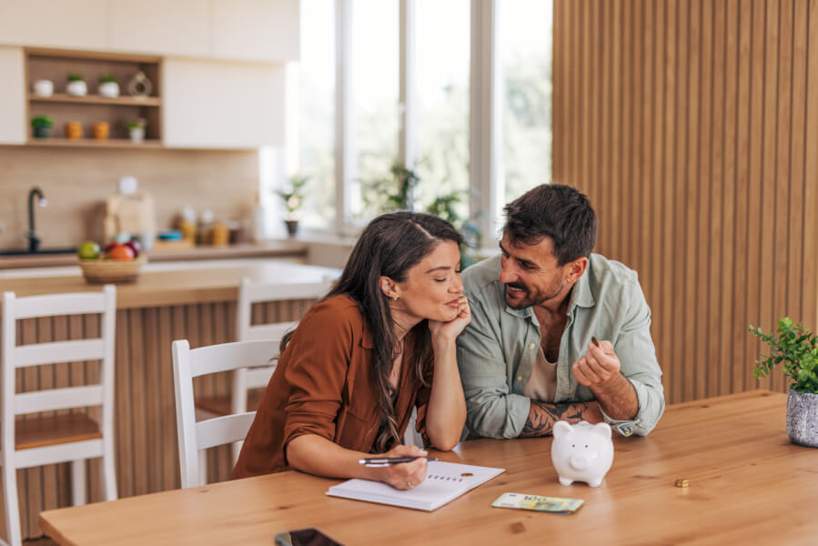 Couple calculant leur taux d’endettement maximum autour d’une table en bois dans une cuisine moderne et lumineuse. La femme, concentrée, prend des notes sur un carnet avec un stylo tandis que l’homme, souriant, explique des détails financiers en tenant des documents et une calculatrice. Sur la table, un petit cochon tirelire blanc et des pièces de monnaie symbolisent l’épargne et la gestion budgétaire. En arrière-plan, une cuisine bien équipée avec des étagères, des plantes et une fenêtre laissant entrer la lumière naturelle. Illustration parfaite pour comprendre comment évaluer son taux d’endettement avant un emprunt immobilier ou un crédit.