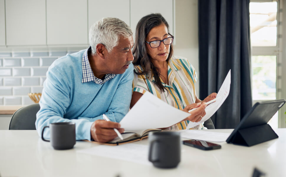 Un couple senior étudiant attentivement des documents fiscaux assis à une table avec des mugs et un tableau numérique, illustrant la préparation du calcul du taux d'imposition pour une location meublée, dans un cadre domestique éclairé et organisé. L'homme, vêtu d'un pull bleu clair, prend des notes dans un carnet tandis que la femme, portant des lunettes et une chemise rayée, consulte des papiers officiels, reflétant une démarche sérieuse de gestion fiscale et de compréhension des obligations liées à la fiscalité locative.