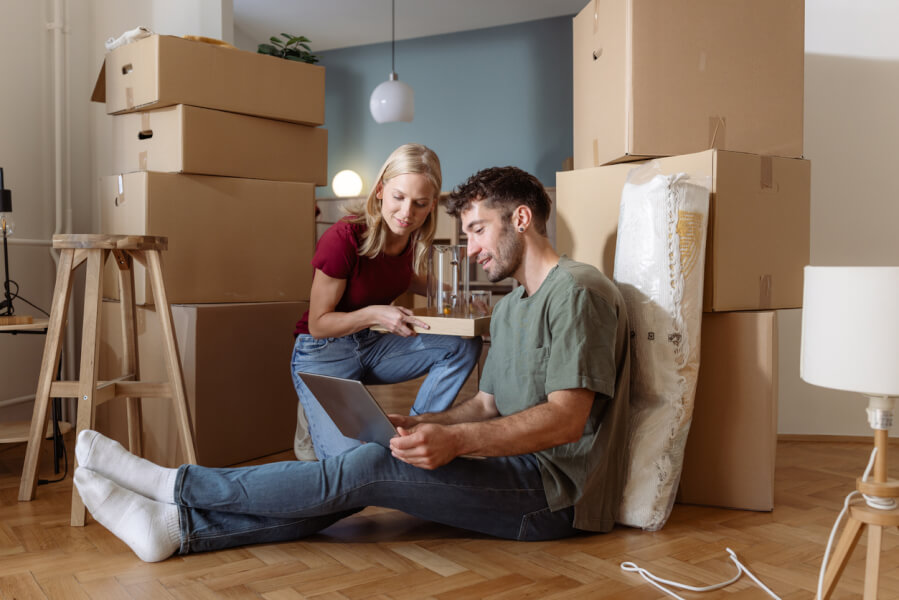 Jeune couple consultant des documents pour réduire la durée d’un préavis de location sur un ordinateur portable dans un salon en cours d’emménagement. Autour d’eux, des cartons de déménagement empilés et des meubles partiellement déballés illustrent une situation de transition locative. L’homme et la femme étudient attentivement des papiers et un écran, probablement à la recherche de solutions légales ou de conseils pour raccourcir leur préavis de location. Cette scène reflète les démarches pratiques et les recherches nécessaires pour optimiser une résiliation de bail en France, tout en mettant en avant l’importance de bien s’informer pour faciliter son déménagement.