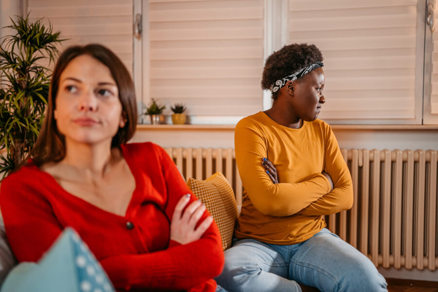 Deux colocataires assises sur un canapé dans un salon lumineux montrant des signes de tension et de désaccord. La femme à gauche, vêtue d’une robe rouge, croise les bras avec une expression faciale sérieuse et légèrement contrariée. À droite, une autre femme portant un haut jaune et un bandana sur la tête, croise également les bras tout en regardant de côté d’un air mécontent. L’arrière-plan montre une pièce bien décorée avec des stores vénitiens, des plantes vertes et un radiateur, illustrant un cadre de vie commun en colocation. Cette image reflète une situation typique de conflit en colocation nécessitant une gestion efficace pour maintenir une bonne entente entre les résidents.
