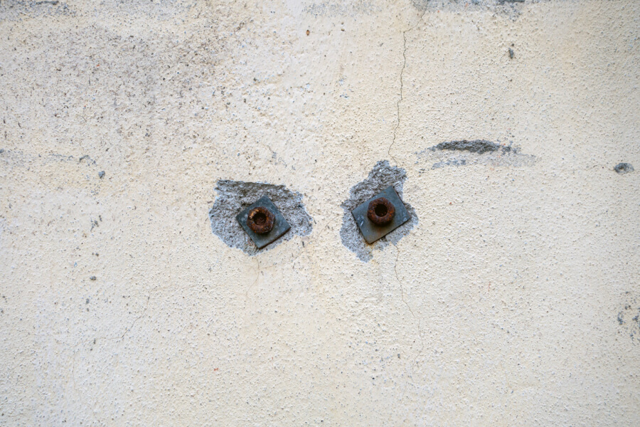 Deux fixations murales rouillées avec écrous encore en place dans un mur enduite de plâtre fissuré et dégradé, illustrant les traces de trous laissés après le retrait d'étagères ou d'éléments fixés au mur lors d'un état des lieux de sortie.