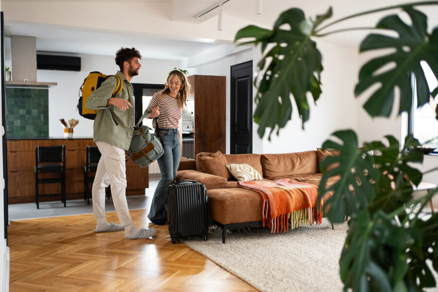 Deux personnes souriantes préparant un voyage dans un appartement meublé lumineux, illustrant l'impact des modifications de la TVA sur les locations touristiques en 2026. L’homme porte un sac à dos jaune et tient une valise, tandis que la femme, avec une valise noire à roulettes, semble discuter joyeusement. Le salon moderne comprend un canapé en velours marron, des coussins décoratifs, une couverture tricotée, une table basse et des plantes d’intérieur. En arrière-plan, une cuisine équipée et des meubles en bois ajoutent au charme de ce logement de vacances, sujet aux nouvelles réglementations fiscales concernant la TVA pour les locations saisonnières.