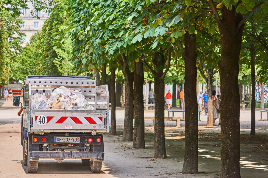 Véhicule utilitaire de collecte des déchets en action dans un parc urbain arboré, chargé de sacs et cartons, avec une signalétique indiquant une limitation de vitesse à 40 km/h, licence française visible GH-237-VD. Scène illustrant la gestion des ordures ménagères en milieu public, avec des passants en arrière-plan, idéale pour évoquer les enjeux de la taxe des ordures ménagères et leur collecte en ville.